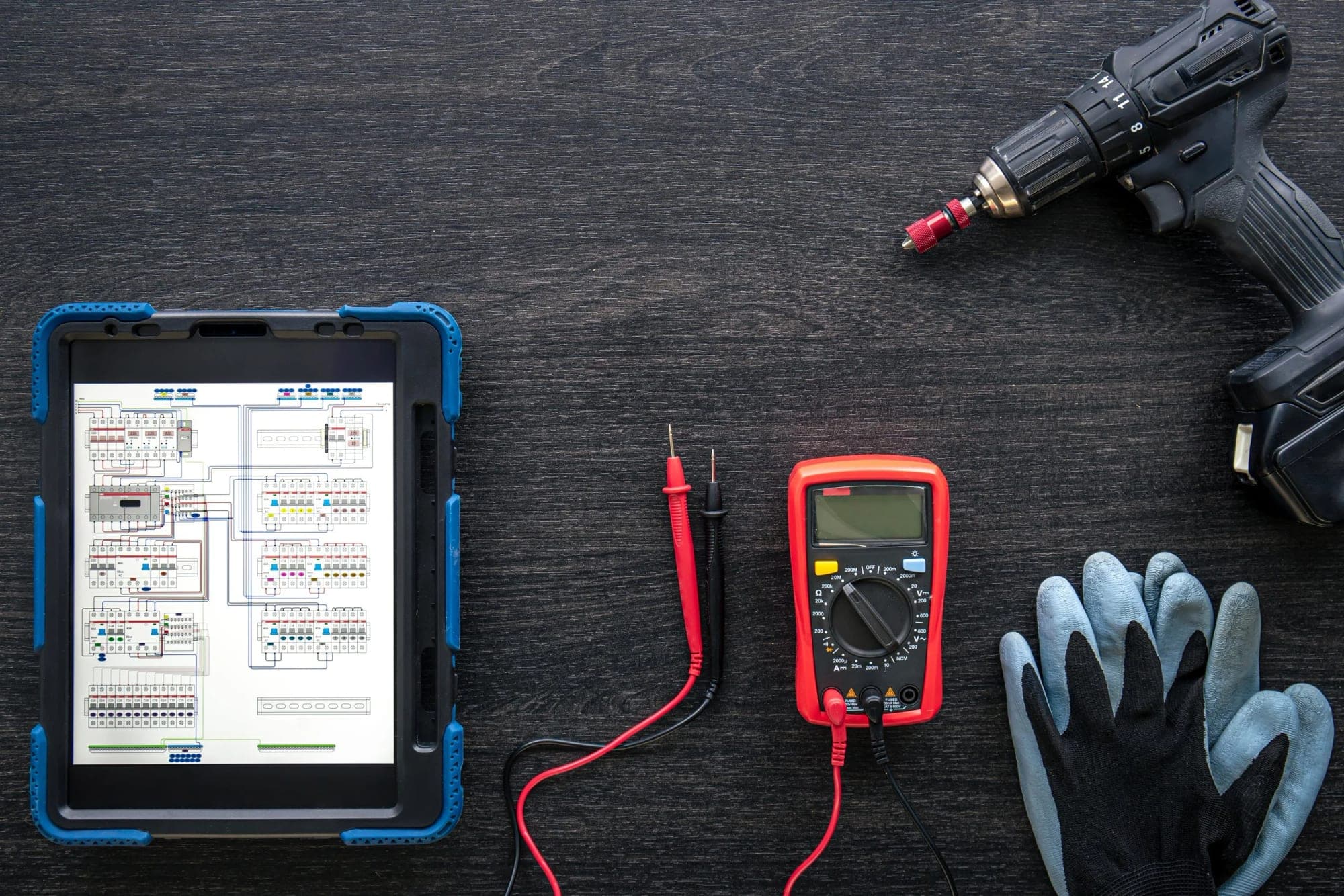 Surfplatta med ett elschema bredvid multimeter, skruvdragare och arbetshandskar